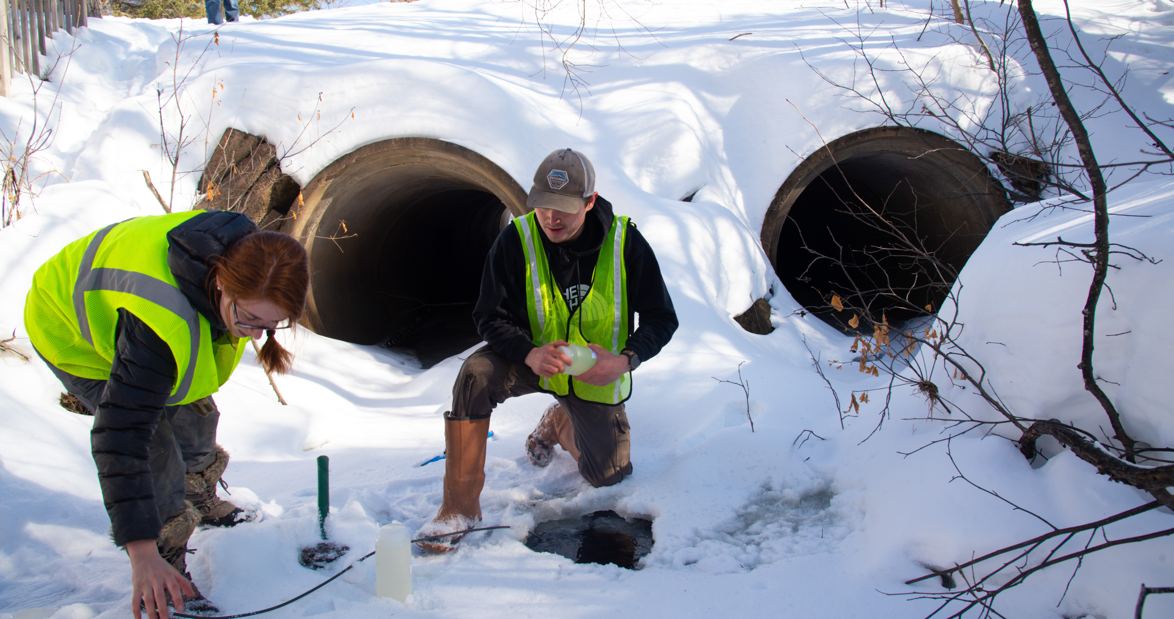 Two students next to a drainage pipe in the snow, using scientific equipment.