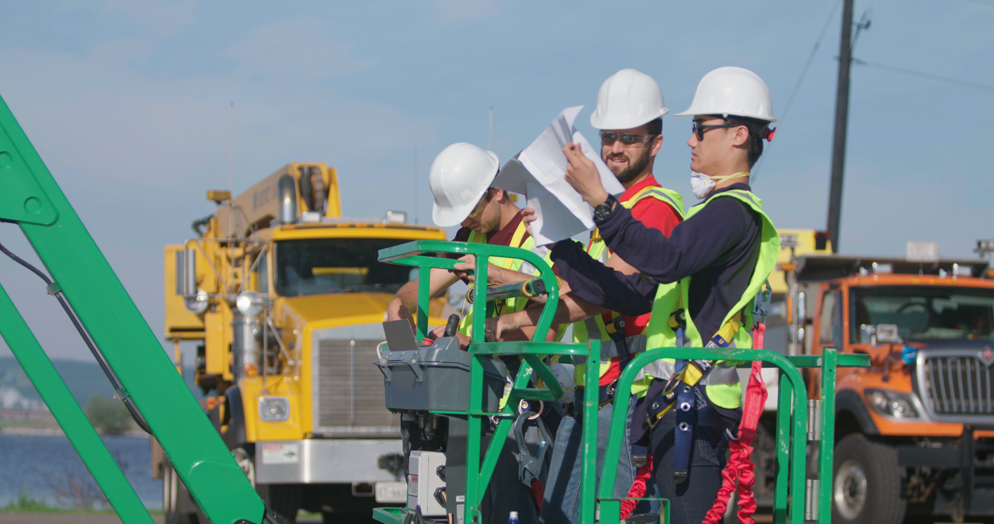 Two men, wearing hard hats and vests, on a construction lift looking at a piece of paper.