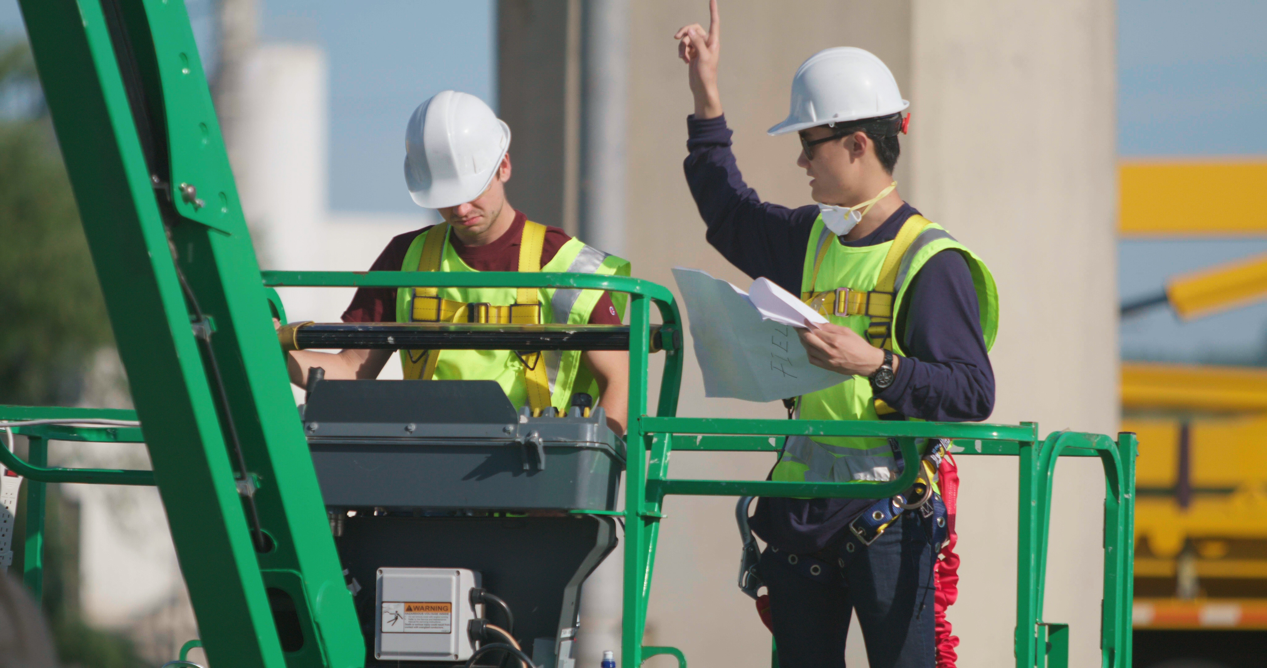 Two men, wearing hard hats and vests, on a construction lift looking at a piece of paper.