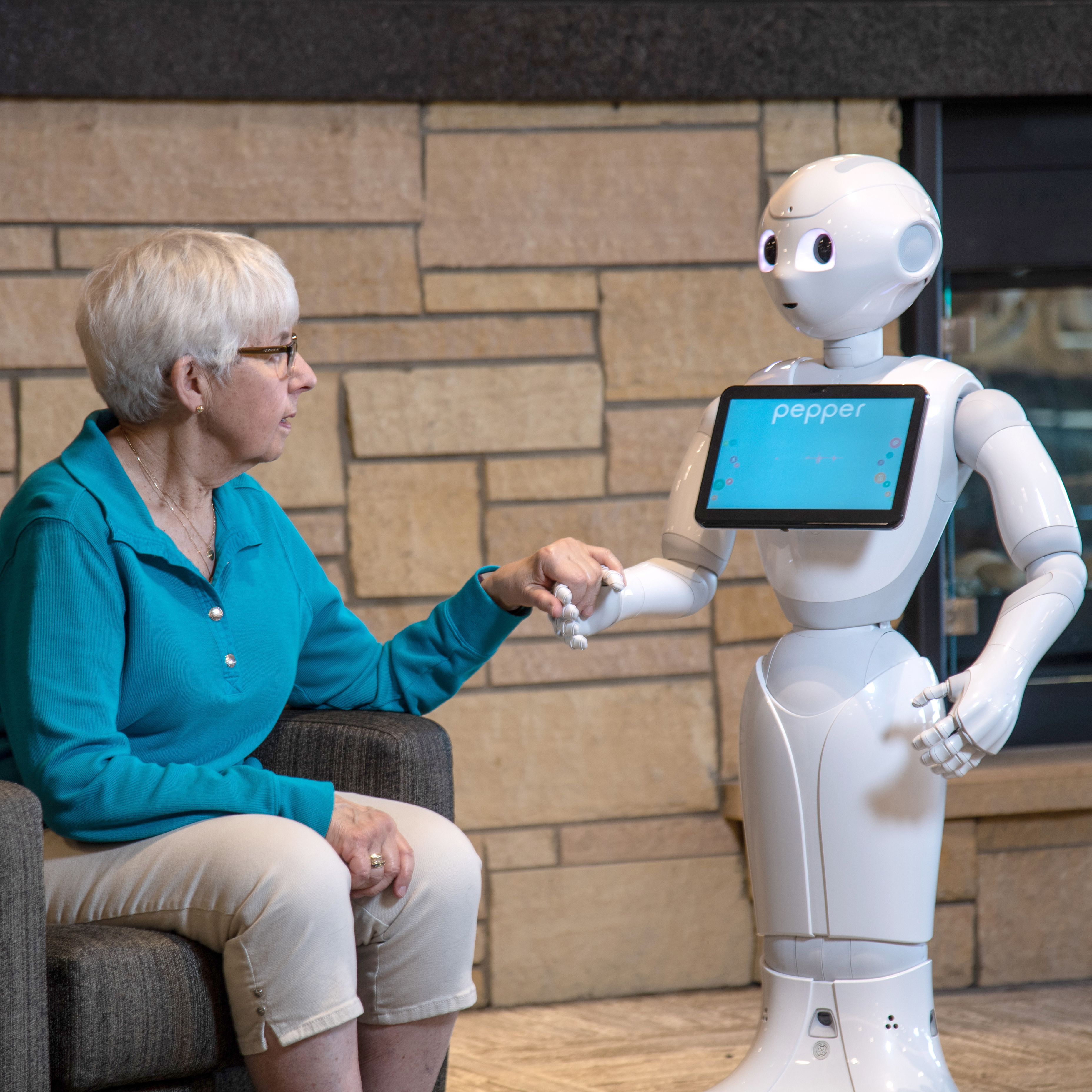 A senior citizen wearing a blue shirt holds hands with Pepper the robot. 