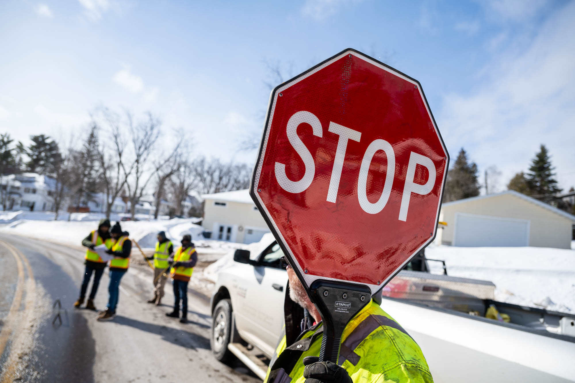 A member of the road testing crew holds a stop sign while others do research in the background on a snowy Duluth street