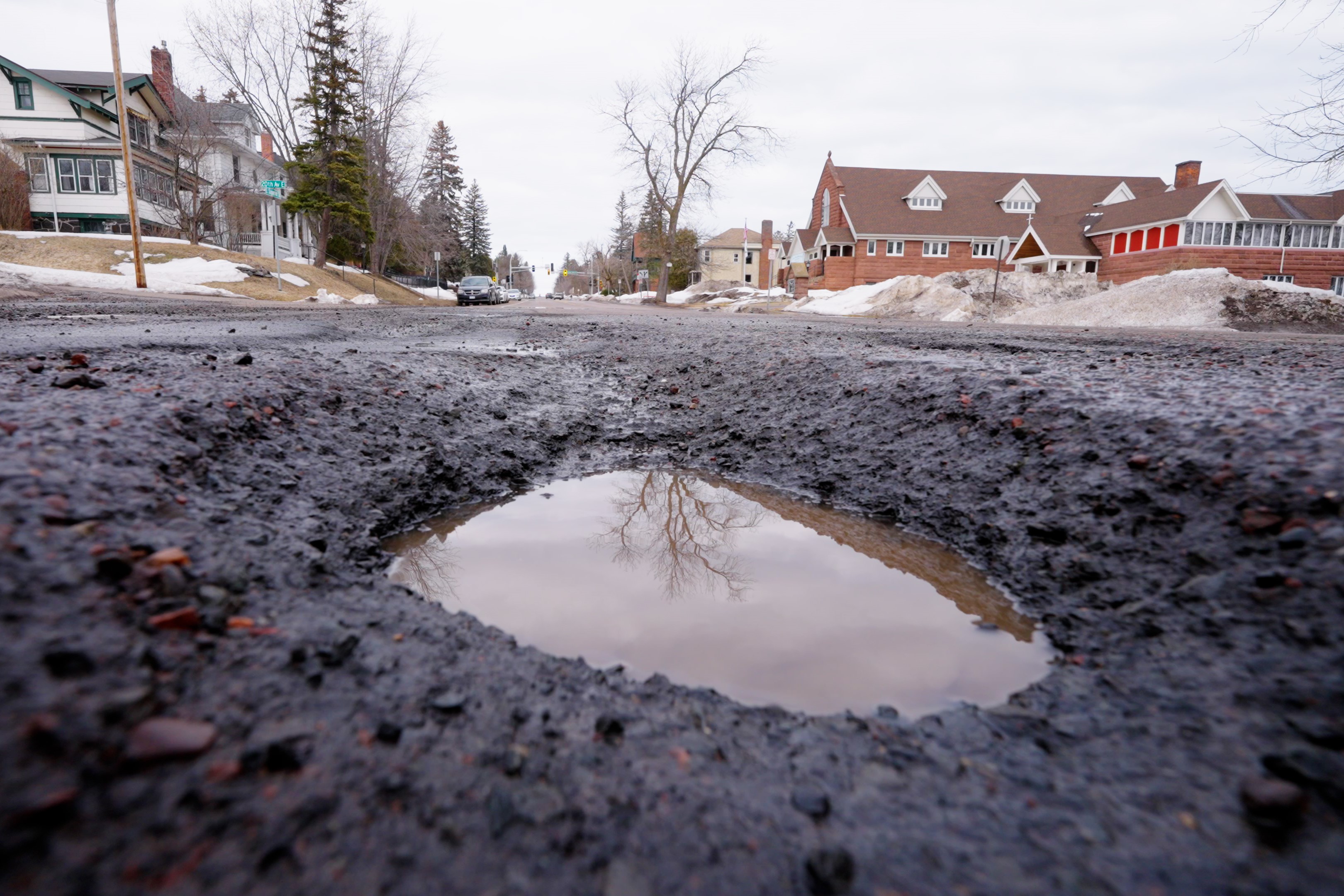 A street-level view of a pothole filled with water with houses and snowbanks in the background
