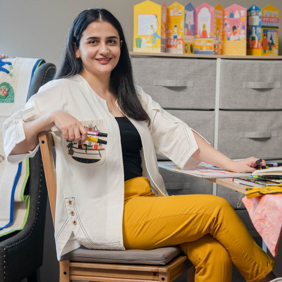 Maryam Khaleghi Yazdi sits at a worktable in her studio, surrounded by her art, while holding markers and smiling at the camera