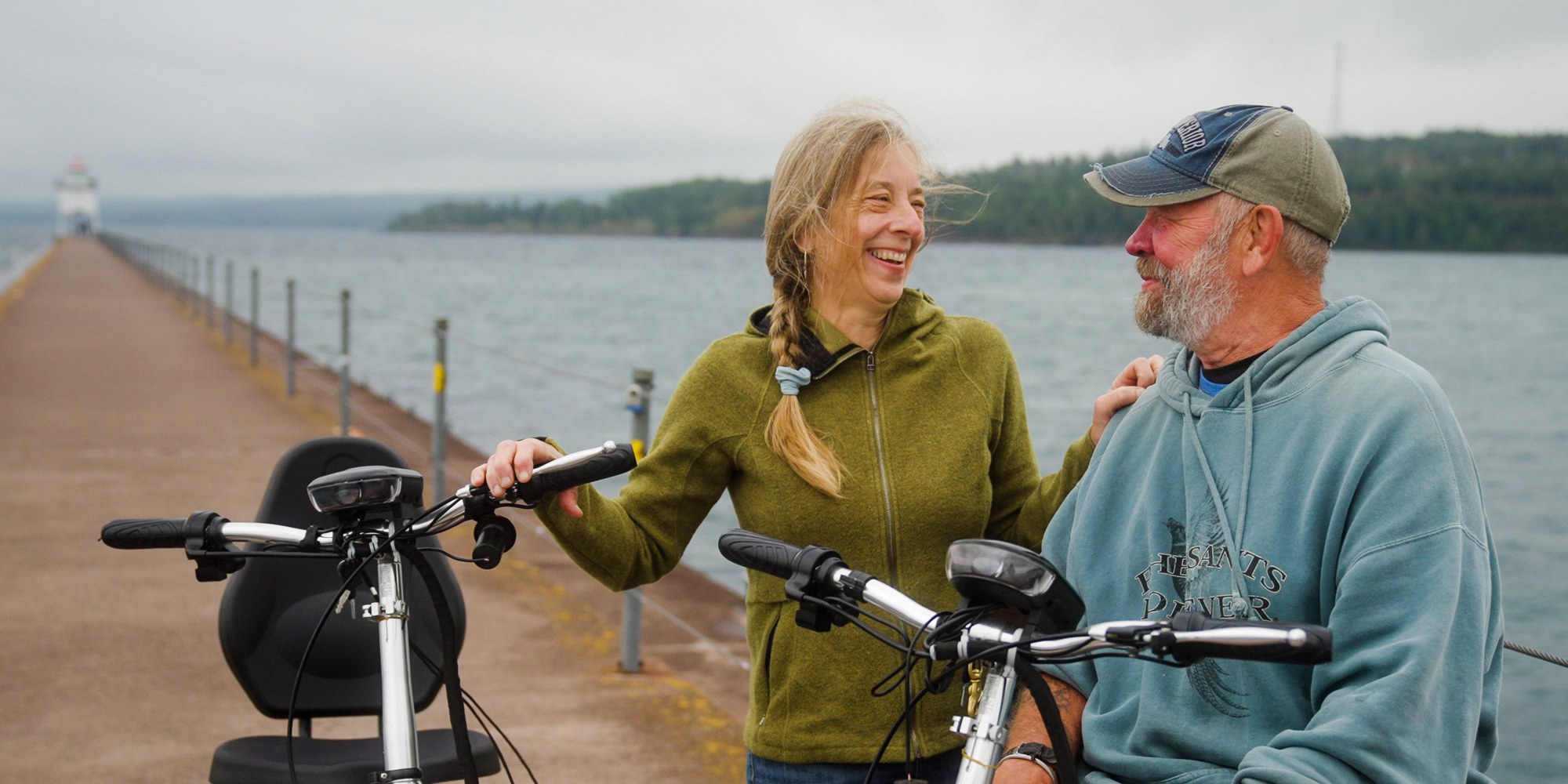 Couple smiling with their bikes along a pier and Lake Superior in background. 