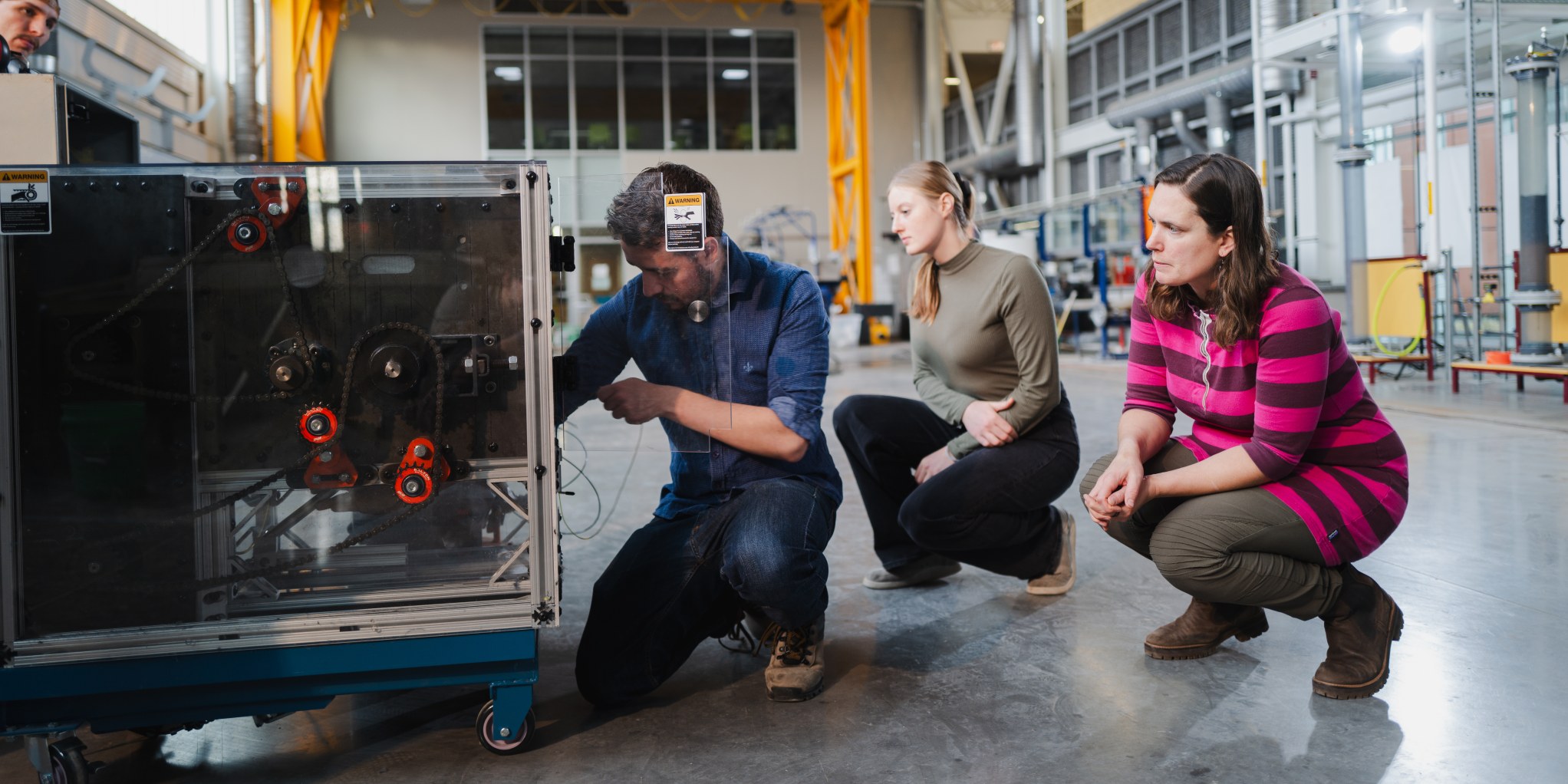 Abbie Clarke-Sather and two UMD graduate students crouch down to inspect the Fiber Shredder machine. 