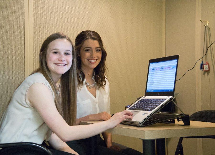 Two young women smiling, sitting next to a laptop.