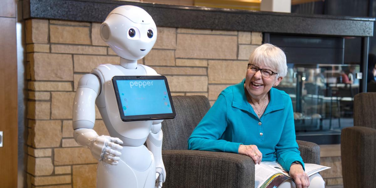 A seated older woman grins past Pepper, a white robot with a large display screen, at someone just off camera